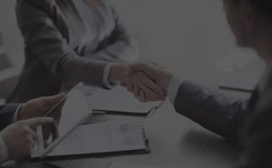 Two people shaking hands at a business meeting with another person looking at a tablet.